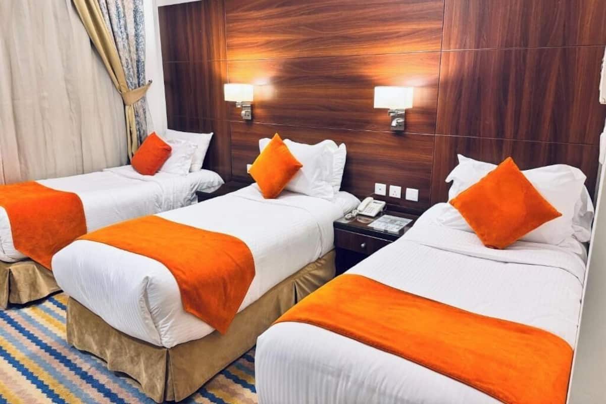 Ein Hotelzimmer mit drei Einzelbetten, jeweils mit weißer Bettwäsche und orangefarbenen Zierkissen. An der Holz-Kopfstütze befinden sich zwei Wandlampen.