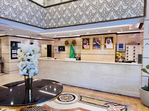 Un área de recepción con piso de mármol, un mostrador de recepción, retratos enmarcados, una bandera y un florero con flores blancas.