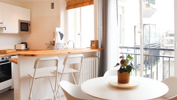 Una cocina moderna con armarios blancos, microondas y tostadora. Hay una zona de comedor con una mesa redonda y sillas, y un balcón con vistas a la ciudad.