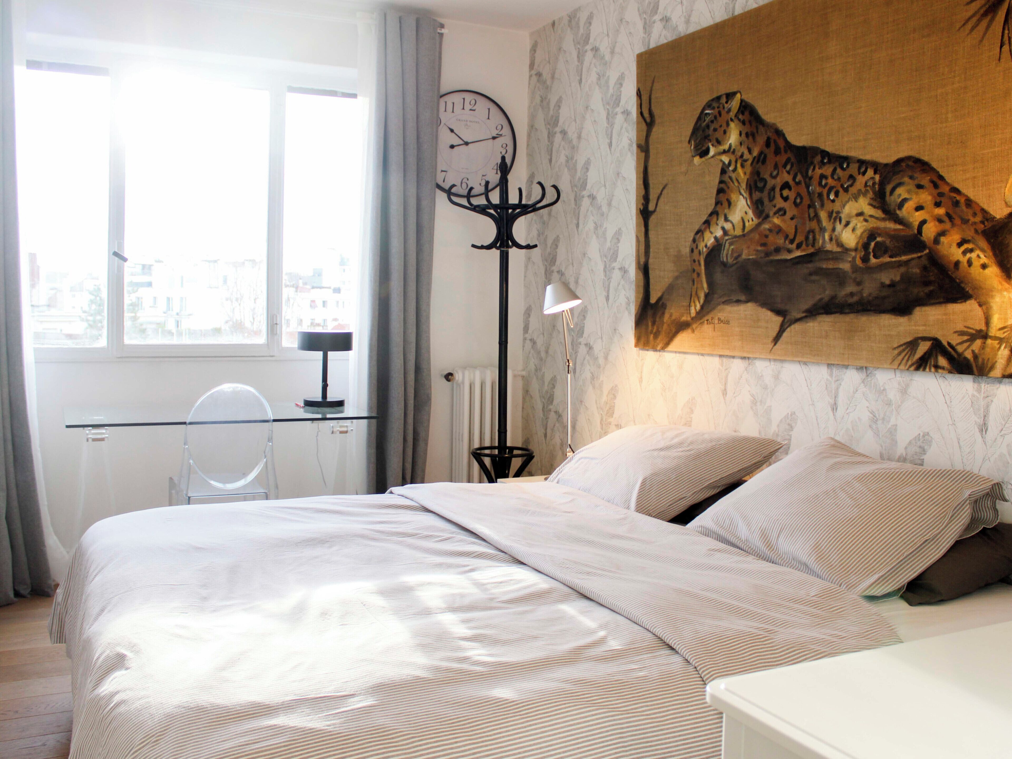 A bedroom with a large bed, a clock, a leopard painting, and a view of the city.