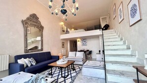 Un'ampia zona giorno con un divano blu, un tavolino, un grande specchio e un lampadario. Si vede una scala che conduce a un piano superiore.