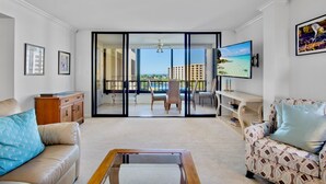 Ein geräumiges Wohnzimmer mit einer großen Schiebeglasdoor, die auf einen Balkon mit Blick auf Strand und Gebäude führt. Es gibt ein Sofa, einen Couchtisch und einen an der Wand montierten Fernseher.