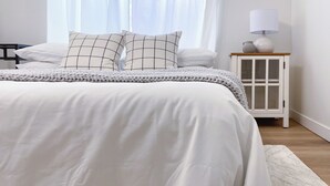 Un lit bien fait, avec une couette blanche et deux oreillers, une table de chevet avec une lampe et un vase, et une fenêtre ornée de rideaux légers.