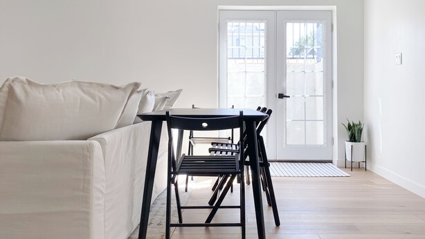 Un salon minimaliste avec un canapé blanc, une table noire et une chaise. On y trouve une porte avec des panneaux de verre et une plante en pot, sur le côté droit.