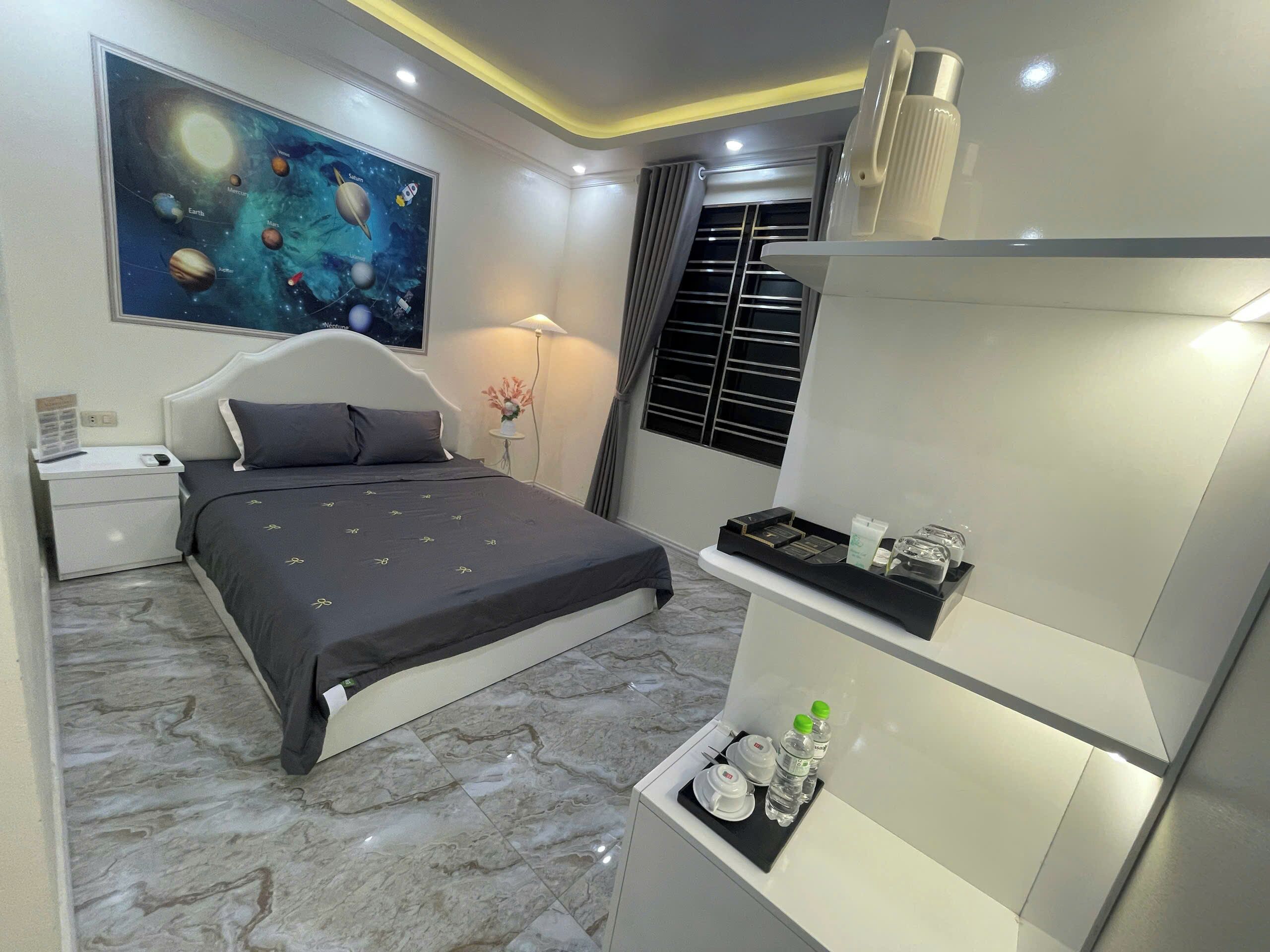 A modern bedroom with a bed, a nightstand, a wall-mounted shelf with toiletries, a painting of planets, and a window with blinds.