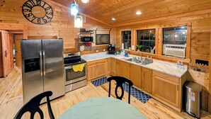 Interior de una cabaña de madera con cocina que incluye un refrigerador de acero inoxidable, horno, microondas y fregadero. Hay un reloj en la pared y una ventana con vistas.