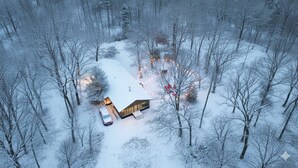Una cabaña nevada con un coche aparcado delante, rodeada de árboles.