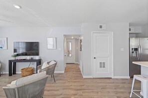 Eine moderne Küche mit einem Edelstahl-Kühlschrank, ein Essbereich mit weißem Tisch und Stühlen sowie ein Wohnbereich mit einer Couch und einem Fernseher.