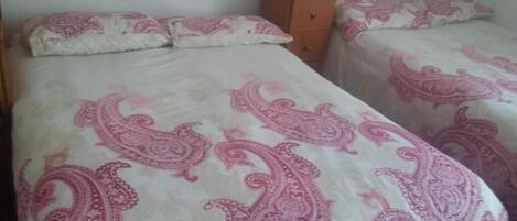 A room with two beds, each with a patterned bedspread. There is a wooden nightstand between the beds.