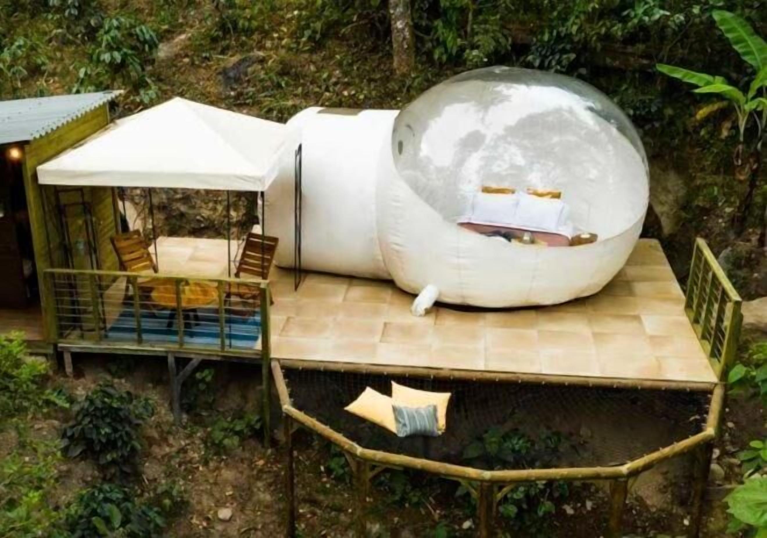 Una estructura esférica con una cama en su interior, rodeada por una terraza de madera y un recinto de vidrio.
