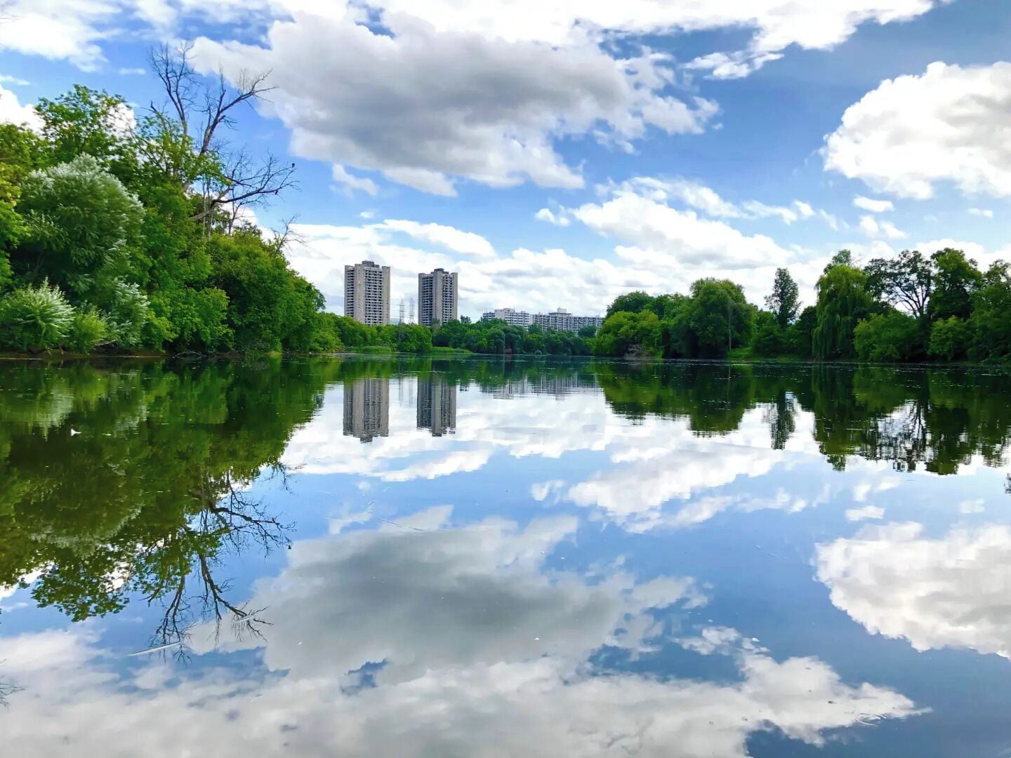 Ein ruhiger See mit Spiegelungen von Bäumen und Gebäuden, umgeben von Grün.