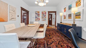 Un espace repas avec une longue table en bois, des chaises blanches, un tapis à motifs et des œuvres d’art accrochées aux murs.