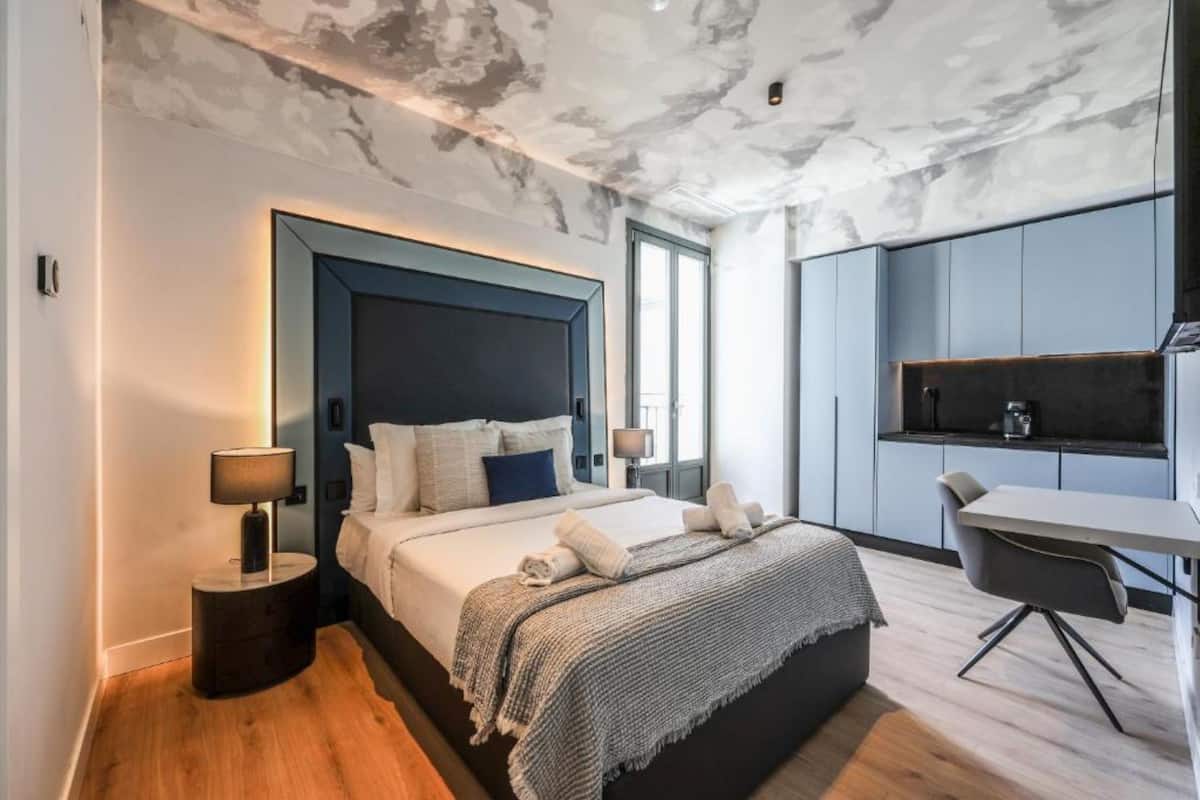 Een moderne slaapkamer met een groot bed, nachtkastjes, een bureau en een kitchenette.