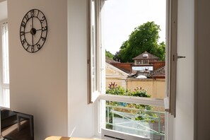 Een kamer met een wandklok, een raam met uitzicht op een huis, en een balkon met een hekwerk.