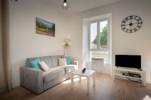 A living room with a grey sofa, a white coffee table, a TV on a stand, a wall clock, and a window with a view of greenery.