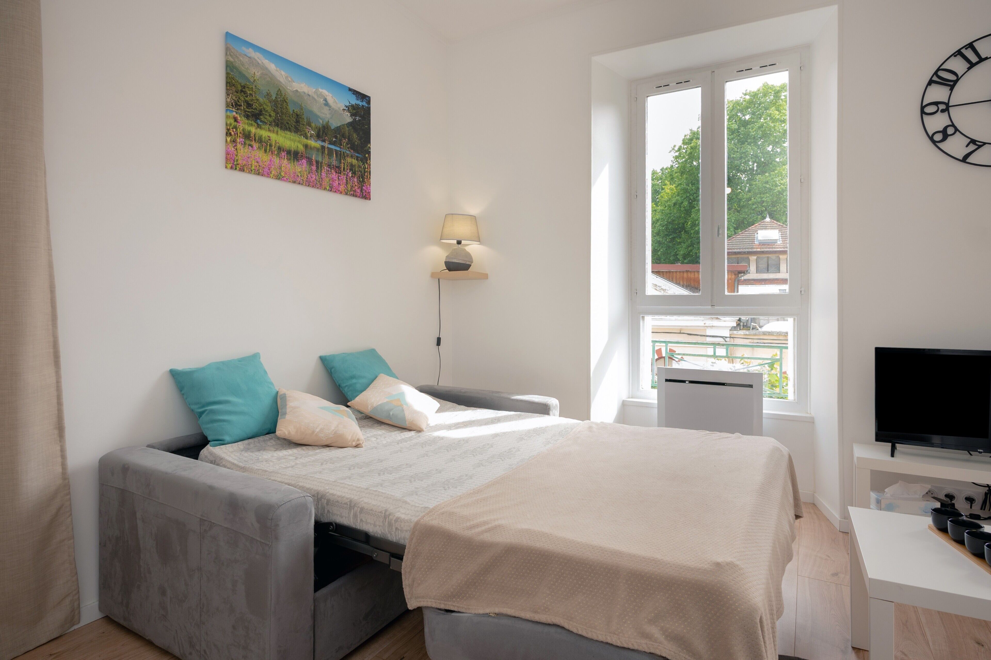 A bedroom with a bed, a desk, a TV, a window, and a painting on the wall.