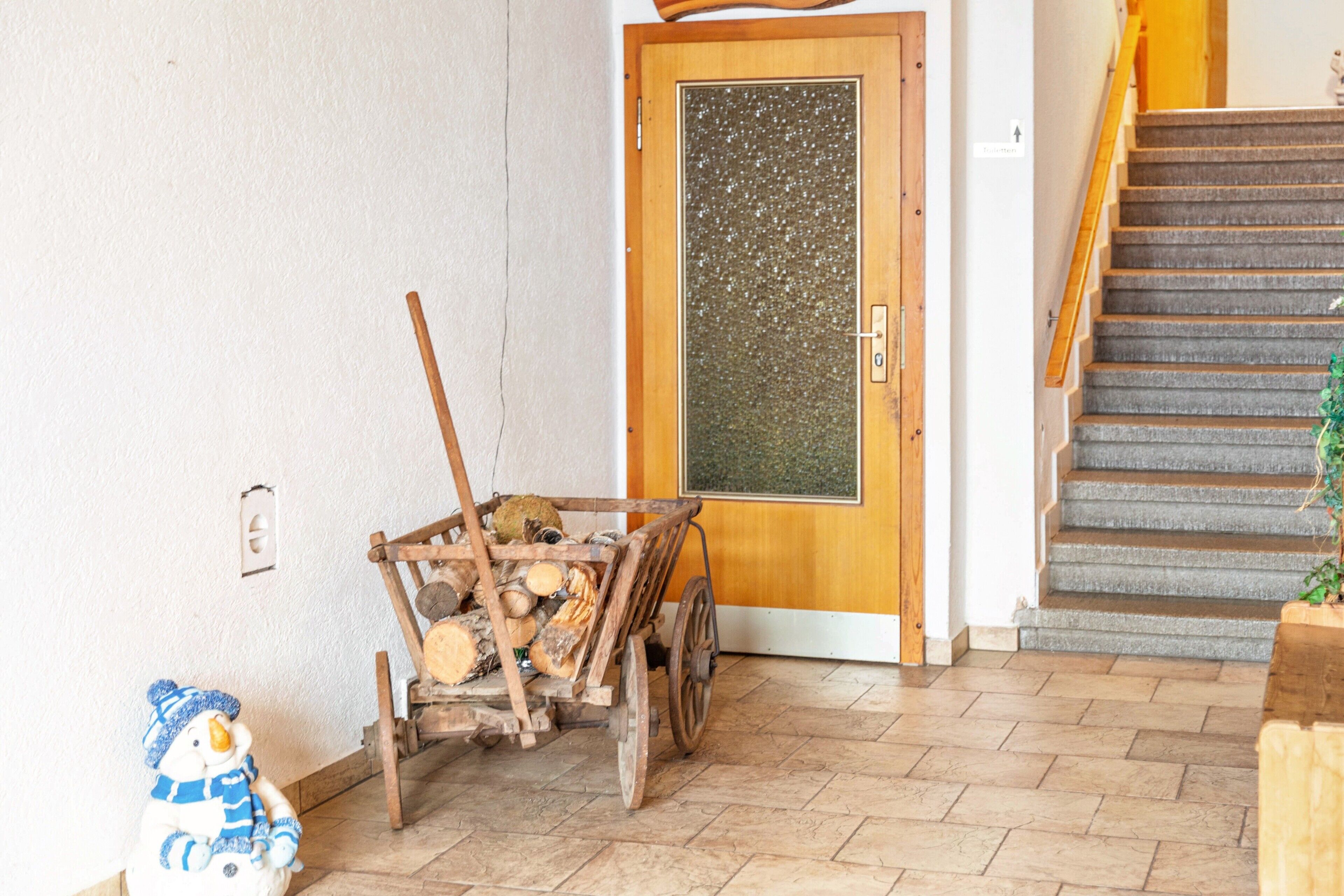 Ein Holzwagen beladen mit Baumstämmen, eine Holztür mit Glasfenster und eine Treppe mit Holzstufen.