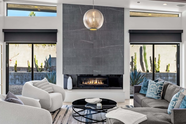 A modern living room with a fireplace, comfortable seating, and large windows overlooking a desert landscape. - Audrey's on Park - Luxury Sunrise Pool Estate (Palm Springs)