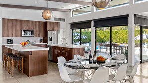 A modern kitchen with a central island, white bar stools, and a dining area with a round table and chairs. - Audrey's on Park - Luxury Sunrise Pool Estate (Palm Springs)