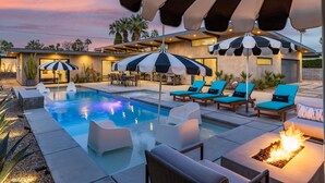 A pool area with lounge chairs, umbrellas, and a fire feature. - Audrey's on Park - Luxury Sunrise Pool Estate (Palm Springs)