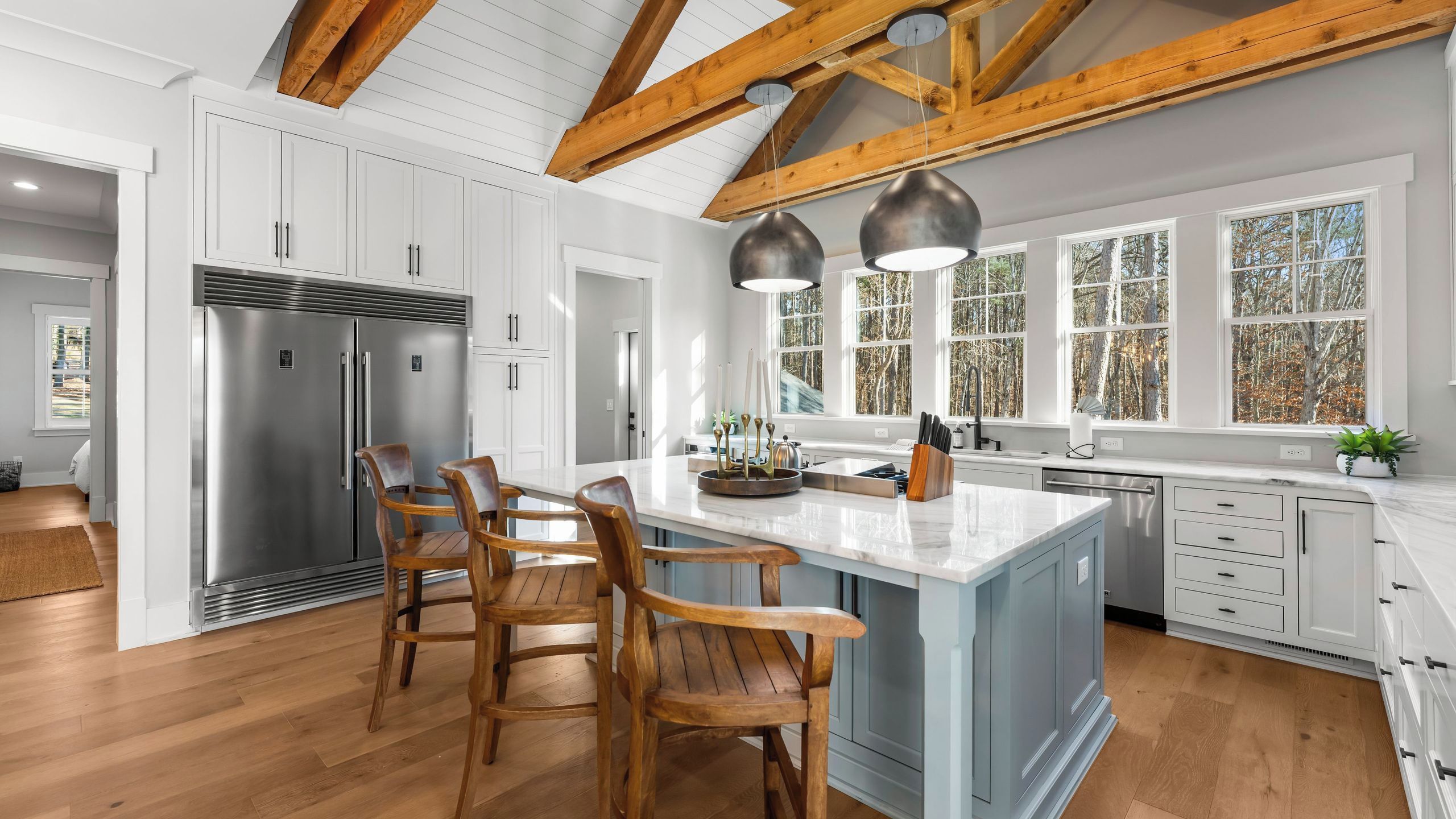 A modern kitchen with a central island, wooden chairs, stainless steel appliances, and large windows.