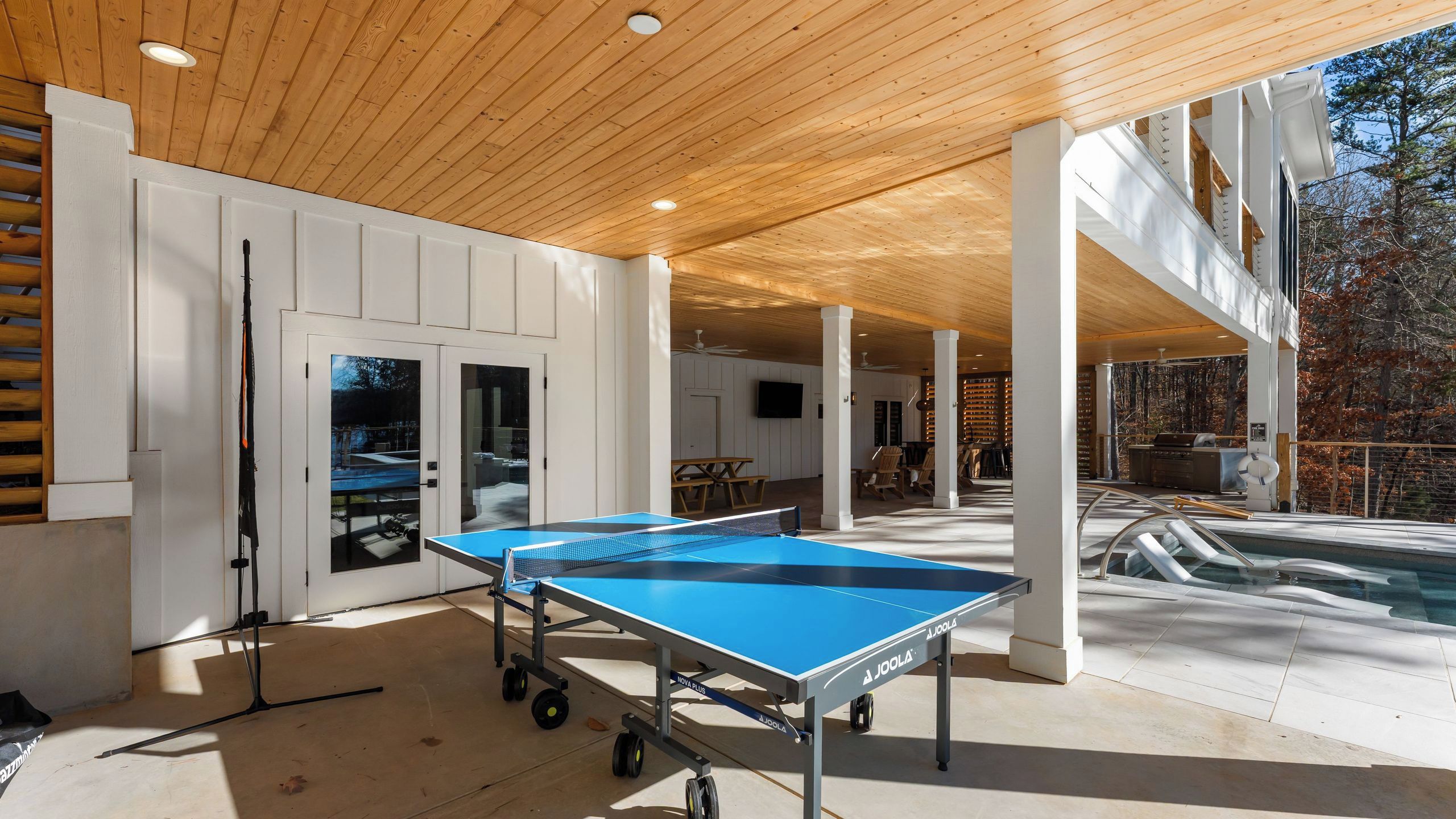 Una mesa de ping pong en el interior, una piscina y un área de estar cubierta con vistas a árboles.