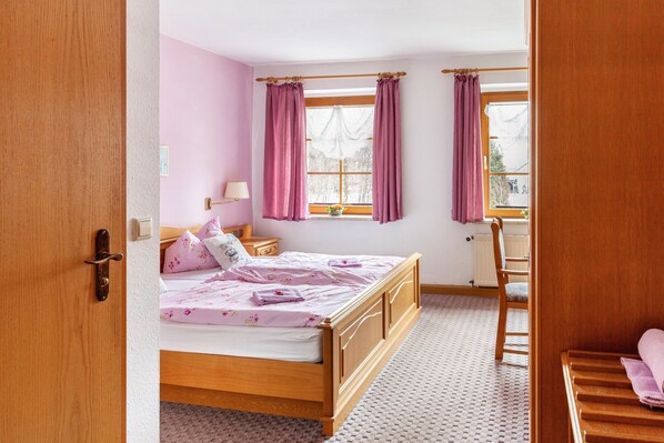Ein Schlafzimmer mit einem hölzernen Bett, einem Fenster mit Vorhängen, einem Nachttisch und einem Sessel.