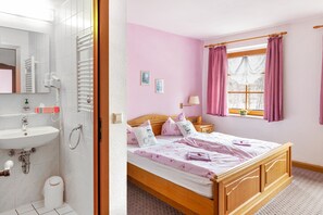 Ein Schlafzimmer mit einem hölzernen Bett, einem Fenster mit Vorhängen, einem Nachttisch und einem Badezimmer mit Waschbecken und Spiegel.