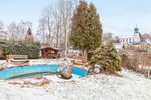 Ein verschneiter Außenbereich mit einer kleinen Holzhütte, einem großen Baum und einem Gebäude mit Gründach im Hintergrund.
