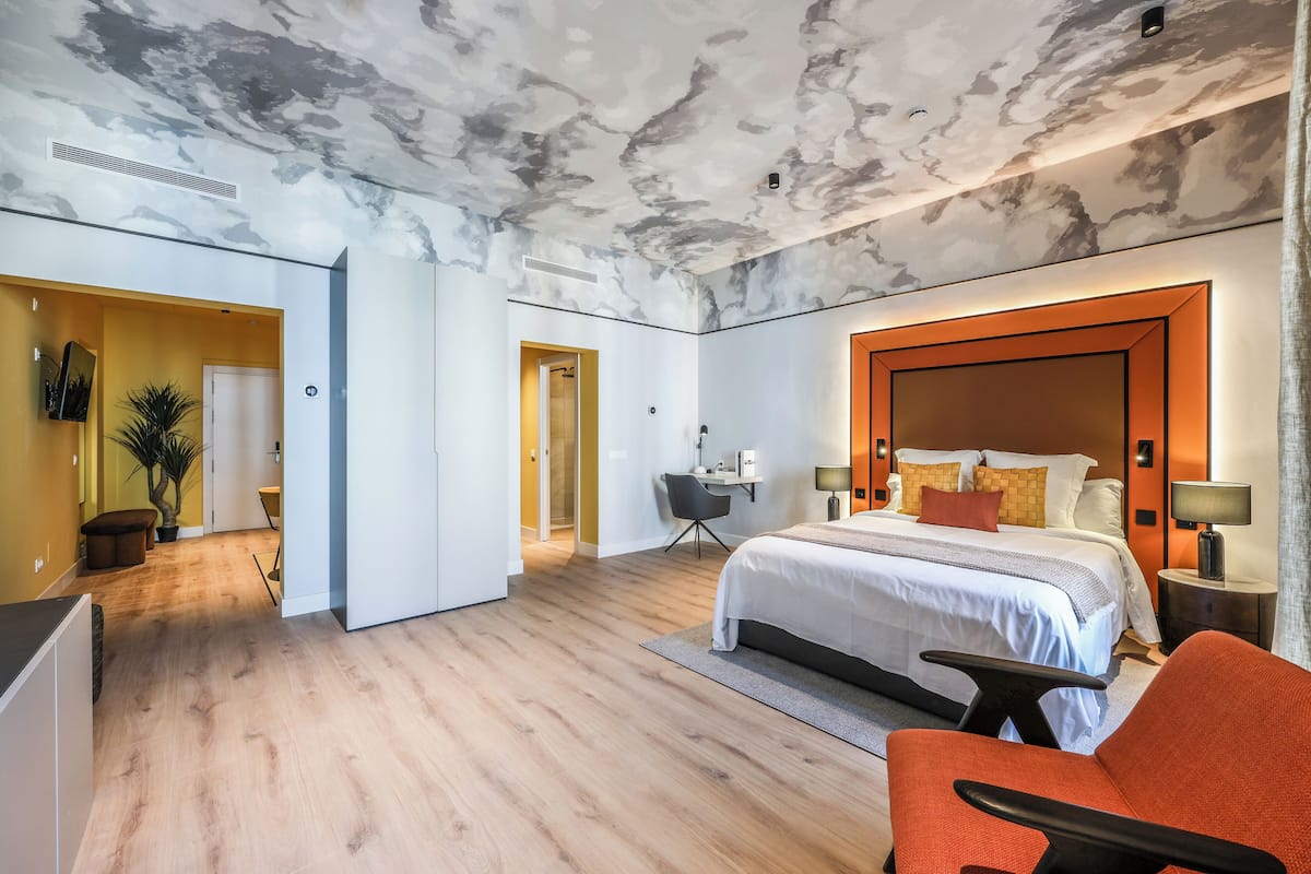 A modern hotel room with a marble ceiling, a large bed with a wooden headboard, a desk with a chair, and a small seating area.