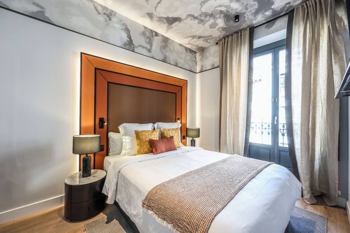 A modern hotel room with a large bed, a wooden headboard, two bedside tables with lamps, and a view of the outdoors through a window with curtains.