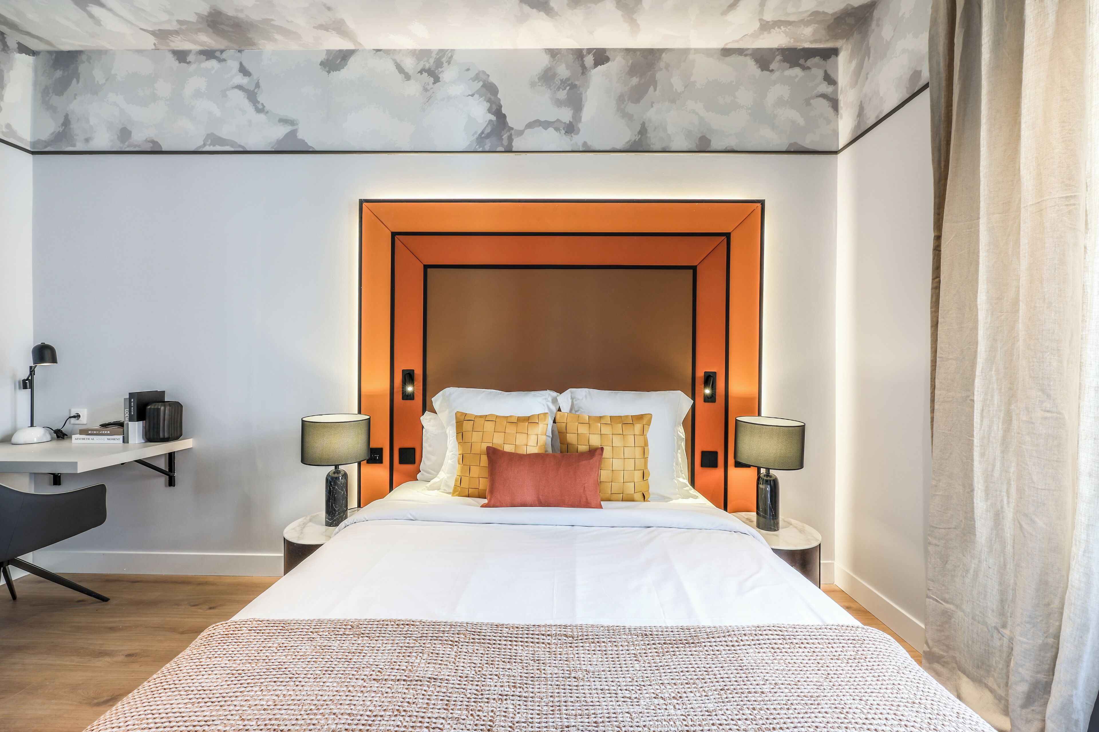A hotel room with a large bed, two bedside tables with lamps, a desk with a chair, and a marble-patterned ceiling.