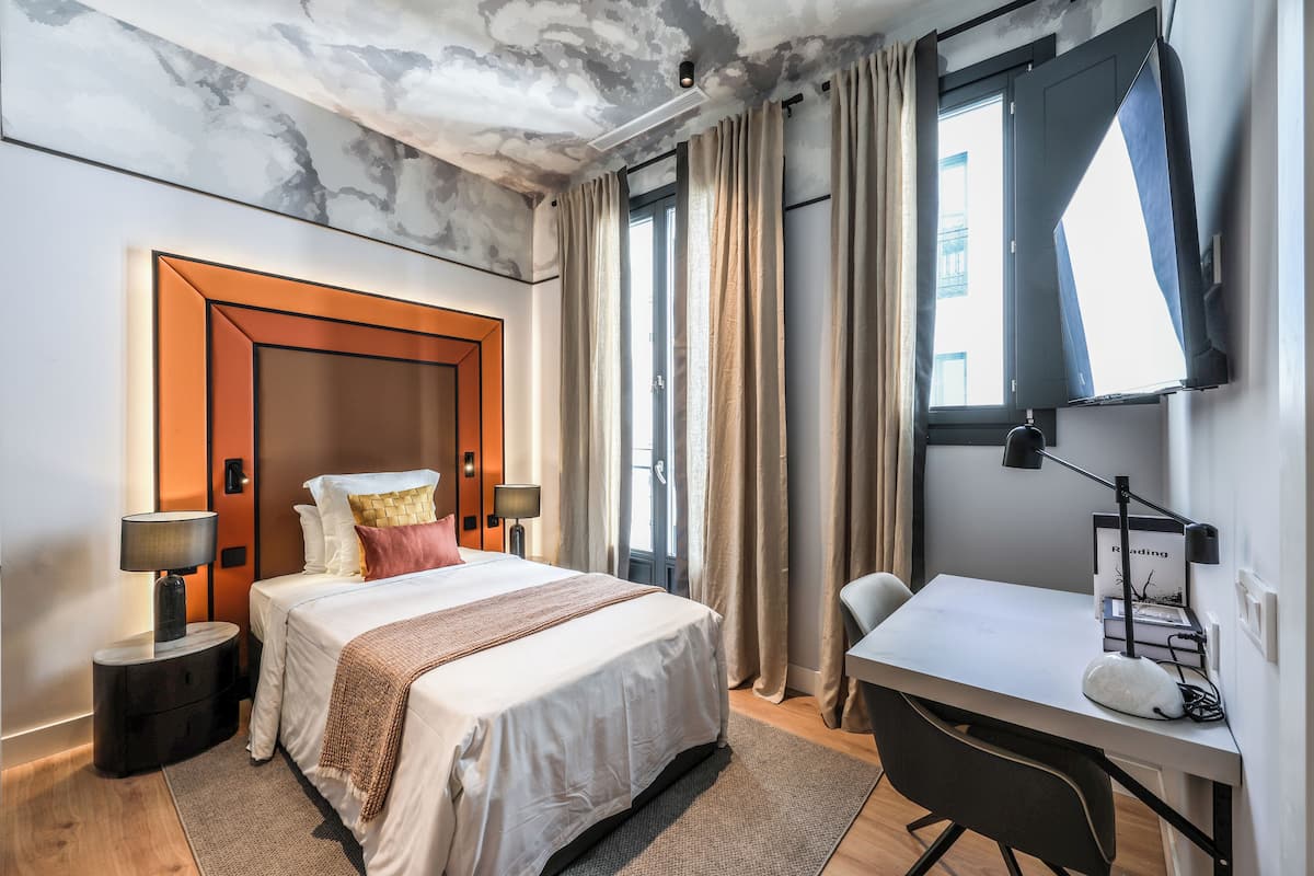 A hotel room with a bed, bedside tables, a desk, a chair, a window with curtains, and a ceiling with a marble-like texture.