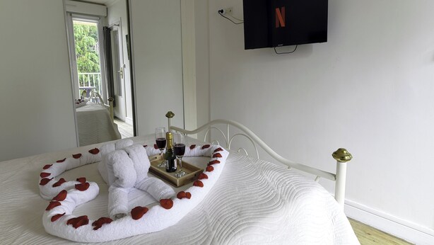 A neatly made bed with rose petals and a tray of wine glasses and bottles, a flat-screen TV on the wall, and a window with a view of greenery. - Le Corot - T2 near city center (Saint-Brieuc)
