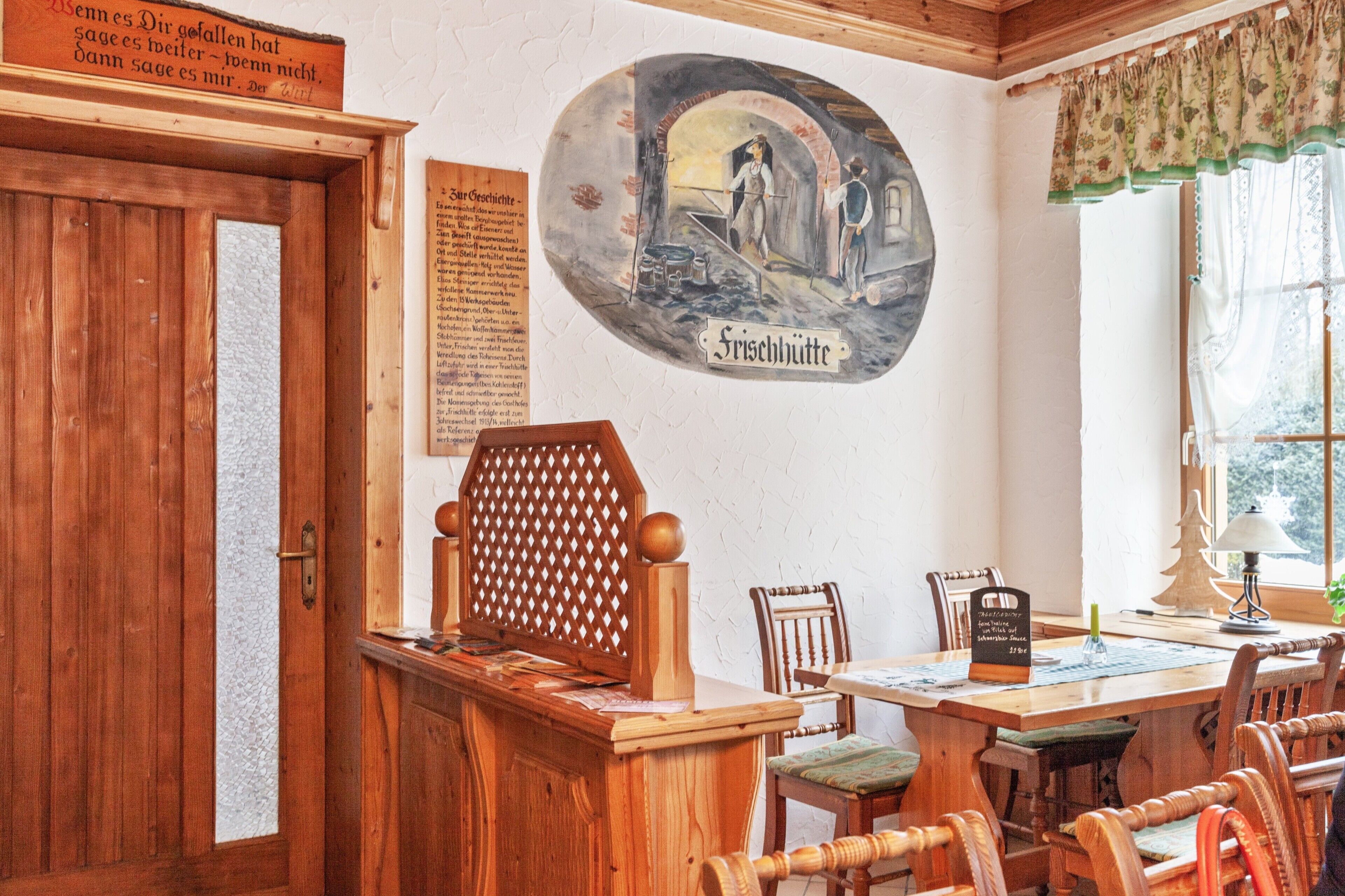 A wooden interior with a dining table, chairs, a wooden desk, and a wall with a mural and text.