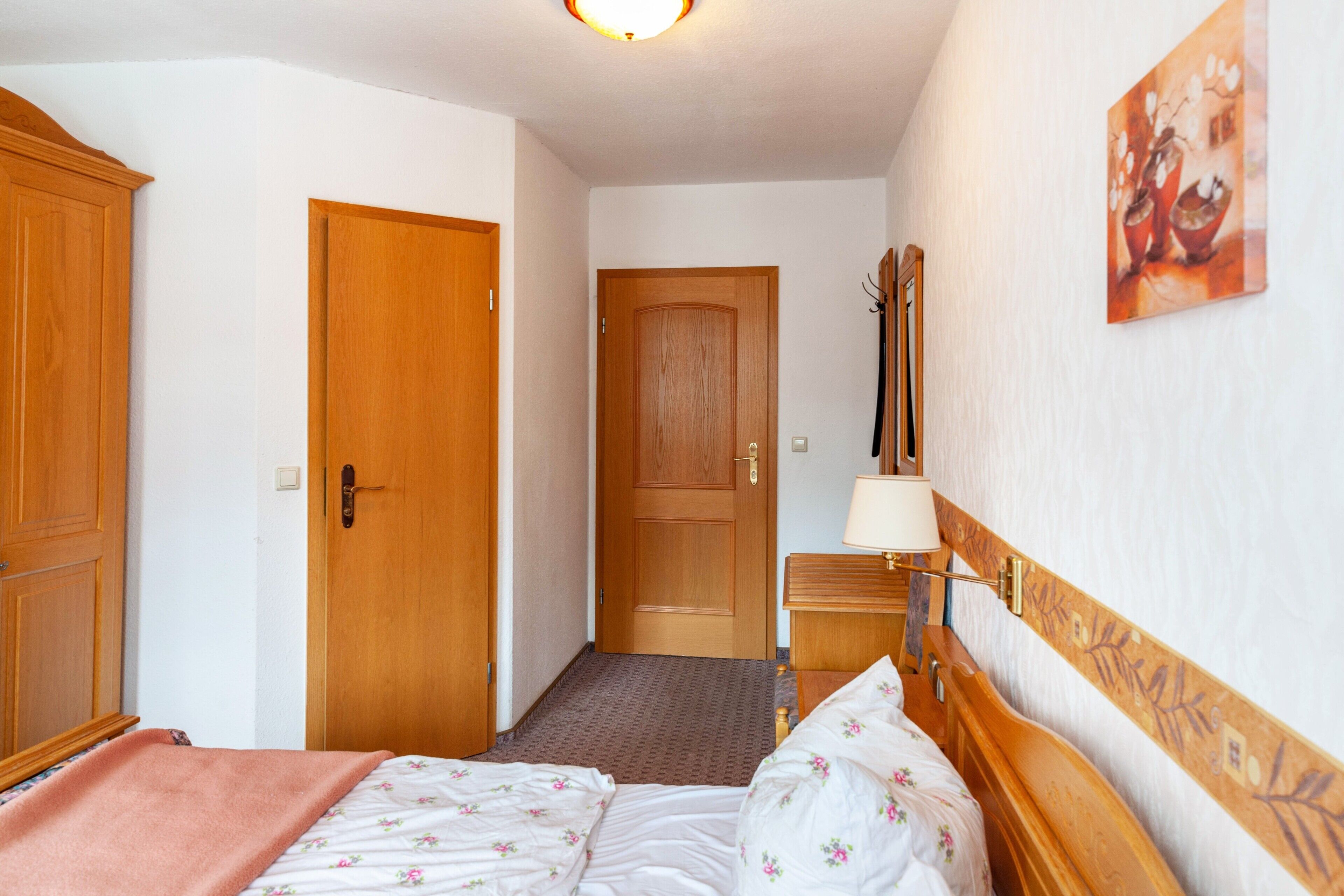 Habitación de hotel con cama, armario de madera, puerta, mesita de noche y lámpara de pared.