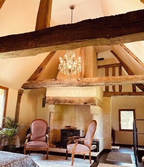 Een rustiek interieur met zichtbare houten balken, een kroonluchter, twee gestreepte fauteuils, een open haard en een raam.