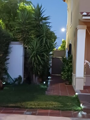 Un chemin menant à un bâtiment doté d’une entrée fermée par un portail, entouré de verdure et de plantes en pot.