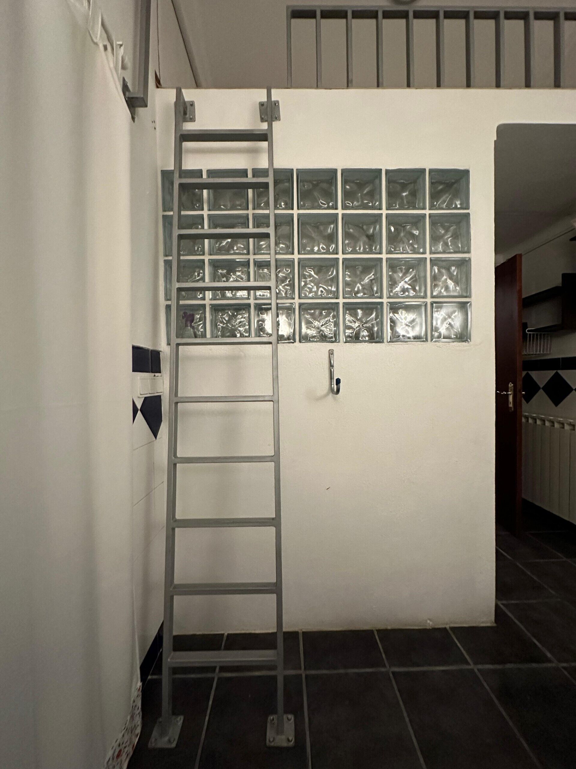 Ein Zimmer mit weißer Wand, einem Fenster mit satinierten Scheiben, einer Metallleiter und einer Tür mit Griff.