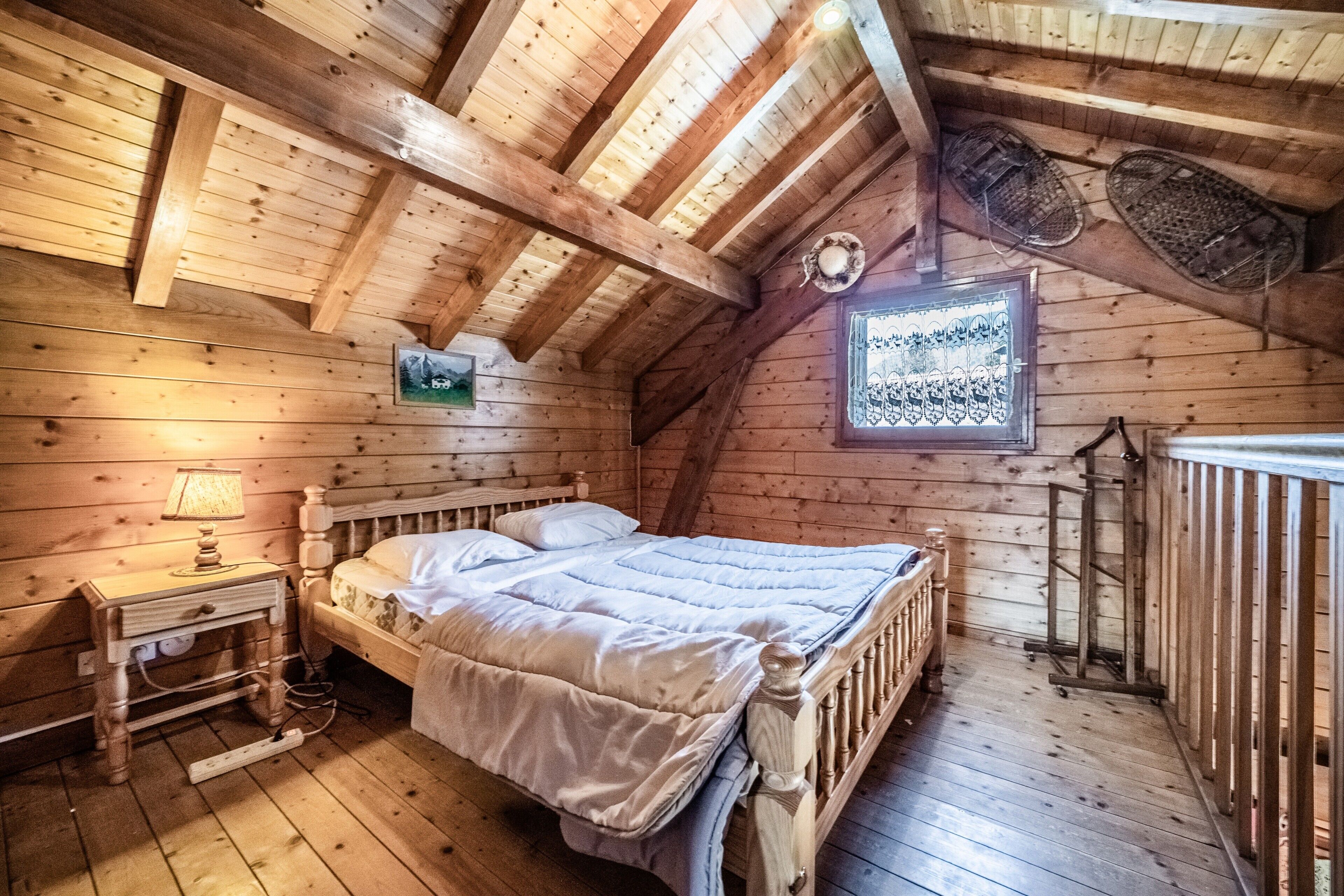 Un dormitorio de cabaña de madera con cama, mesita de noche y ventana con detalles decorativos.