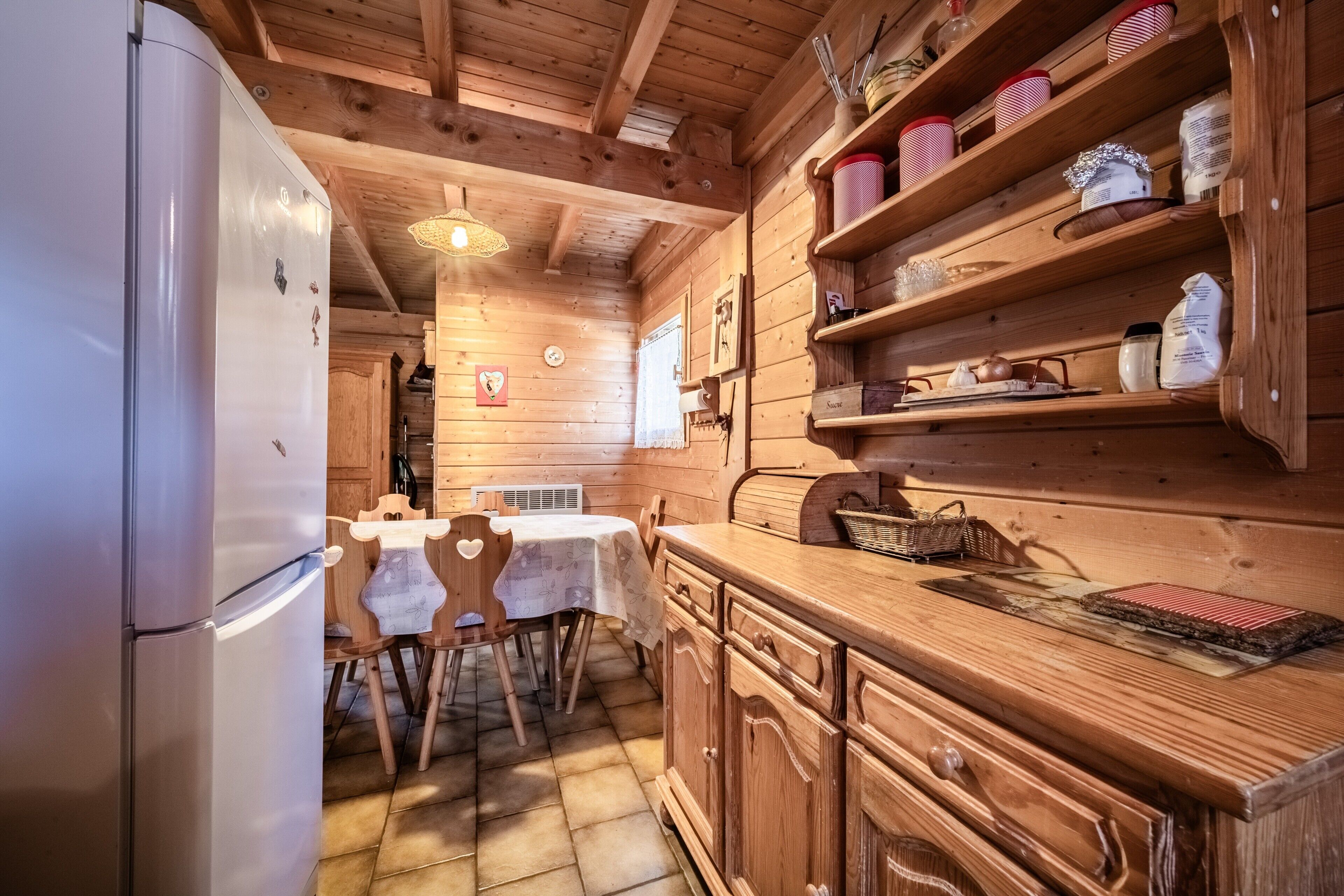 A wooden cabin interior with a refrigerator, wooden dining table, chairs, and shelves with jars and baskets.