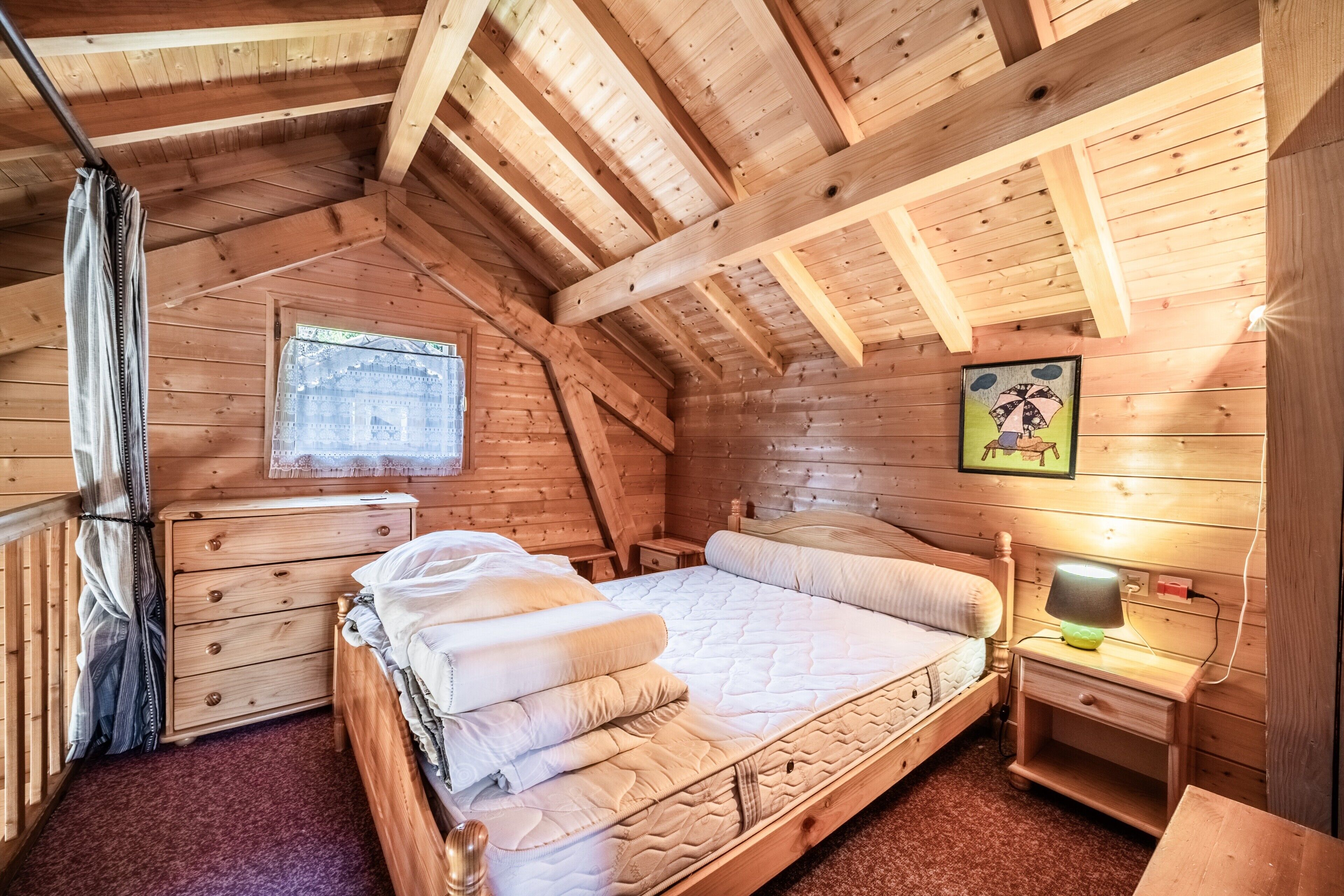 A wooden cabin bedroom with a lofted ceiling, a bed, a nightstand, a dresser, and a window.