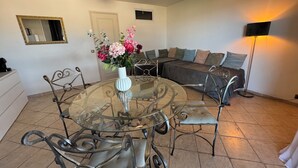 Un comedor con mesa de vidrio y sillas de metal, un sofá con cojines, un florero con flores y una lámpara de pie.