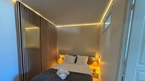 Una cama bien hecha con un edredón oscuro, dos lámparas de noche, un panel de madera y un espejo.