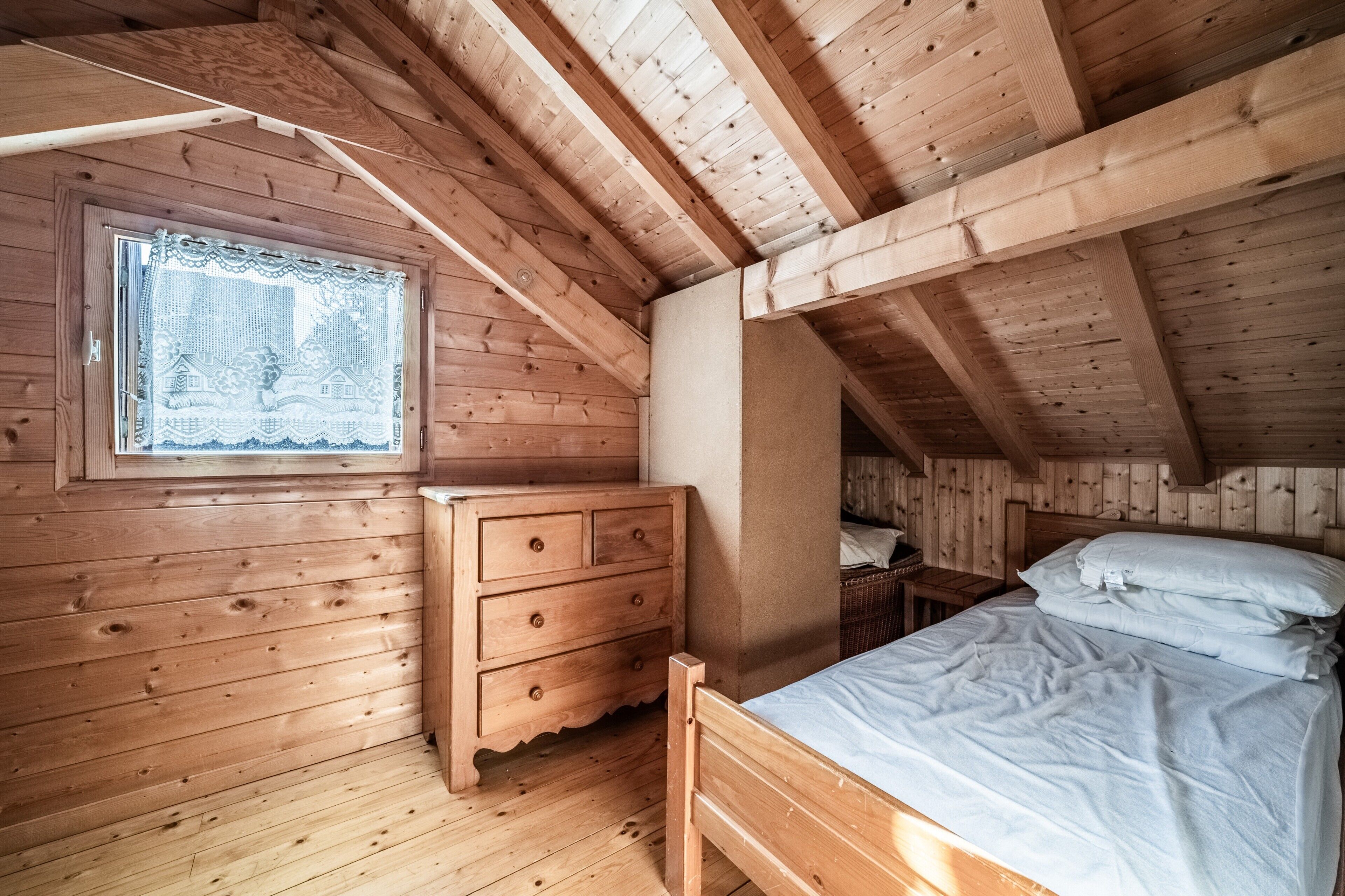 A wooden cabin bedroom with a bed, dresser, and window with lace curtain.