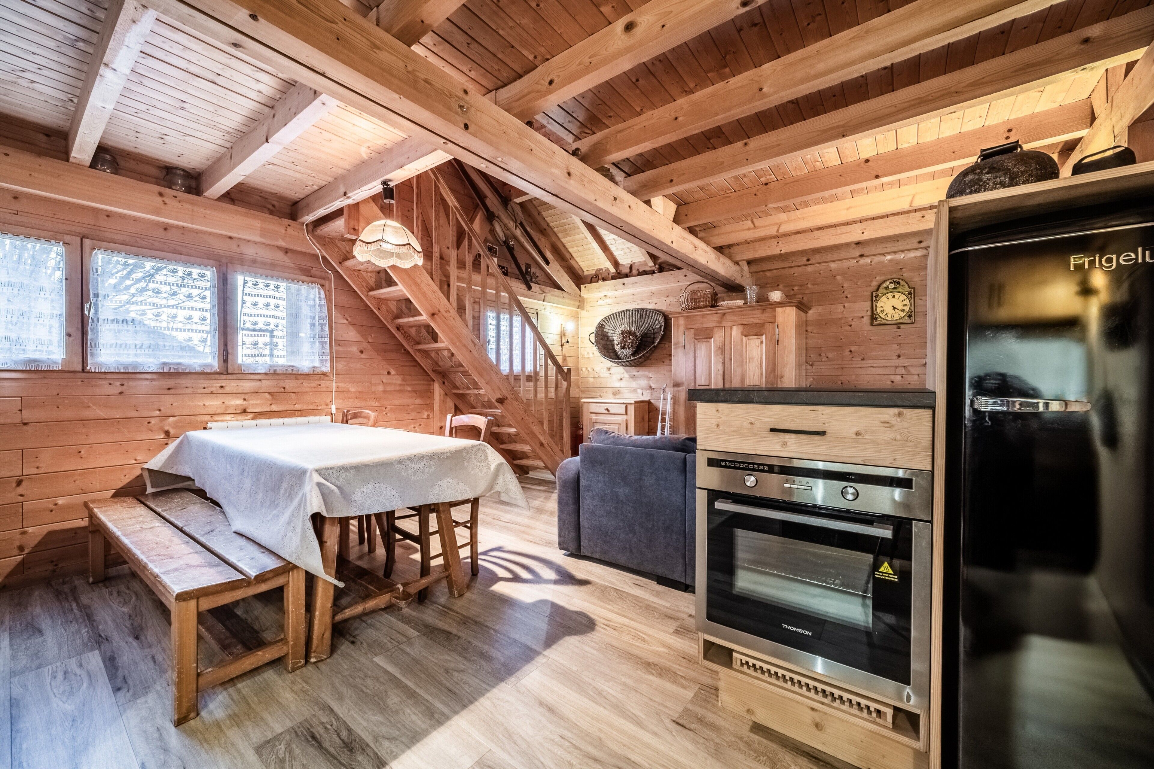 A wooden cabin interior with a dining table, bench, staircase, and a kitchen area with a refrigerator and oven.