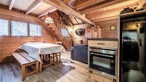 A wooden cabin interior with a dining table, bench, staircase, and a kitchen area with a refrigerator and oven. - Chalet “4p Comfort - Les Trolles”: Mountain View, Private Terrace & Wi-Fi (Verchaix)