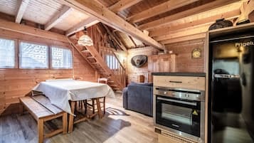 A wooden cabin interior with a dining table, bench, staircase, and a kitchen area with a refrigerator and oven.