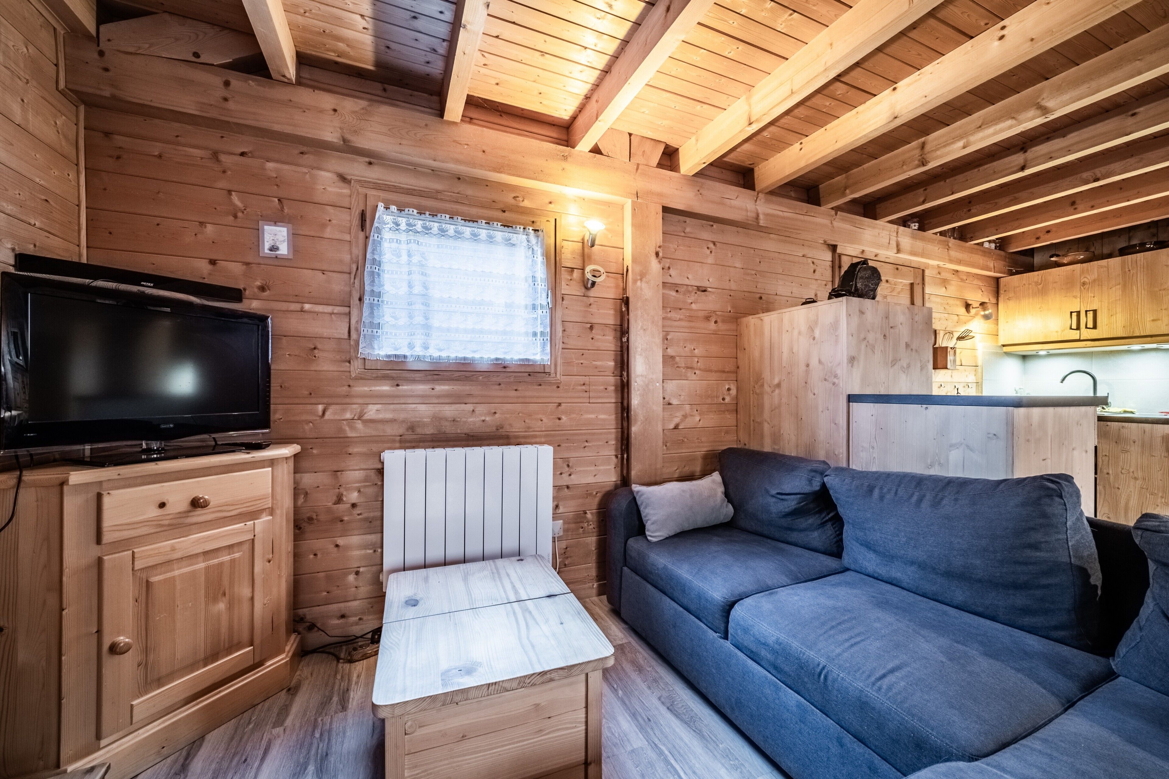 A wooden cabin interior with a television, a sofa, a small table, and a window with curtains.