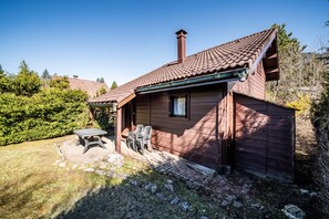 Puinen mökki, jossa on tiilikatto, savupiippu, terassi pöytineen ja tuoleineen sekä kivestä tehty polku.